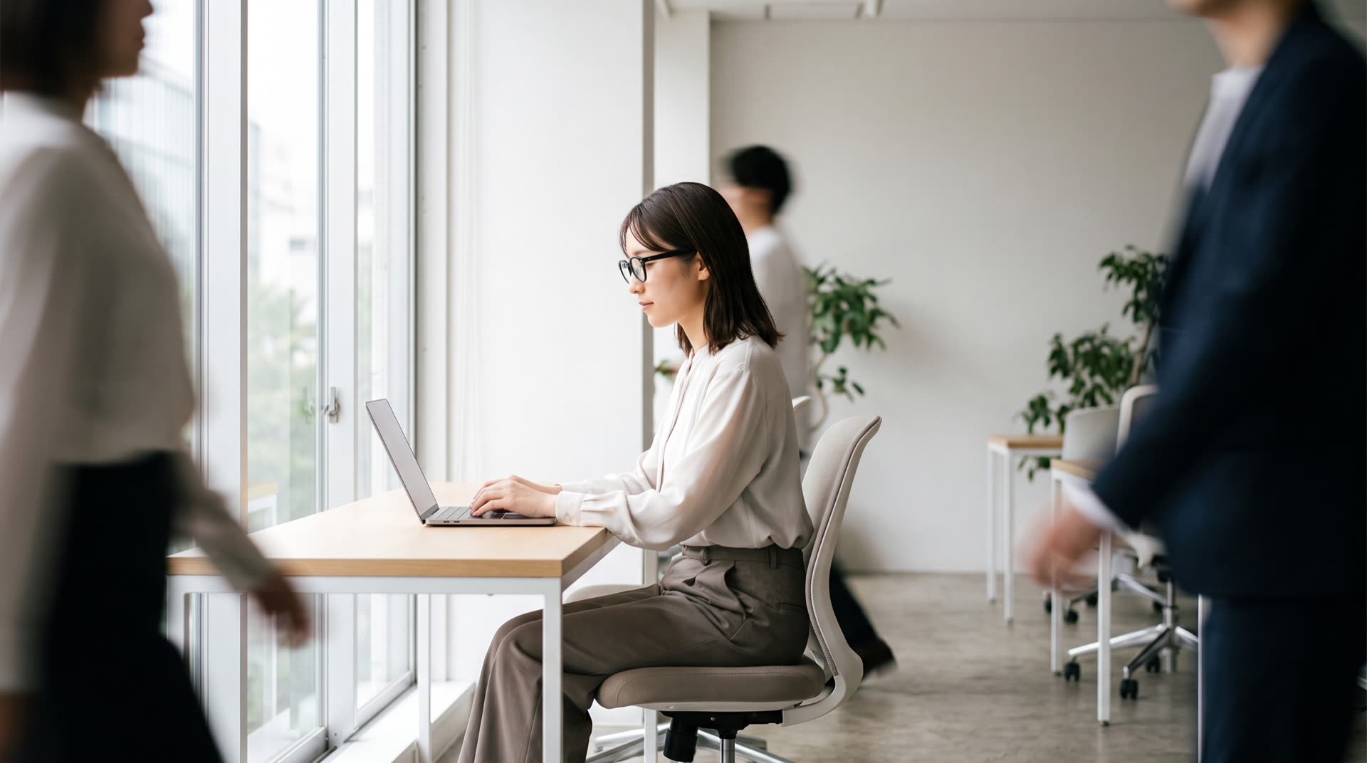 オフィスの窓際でノートPCに向かうメガネの女性
